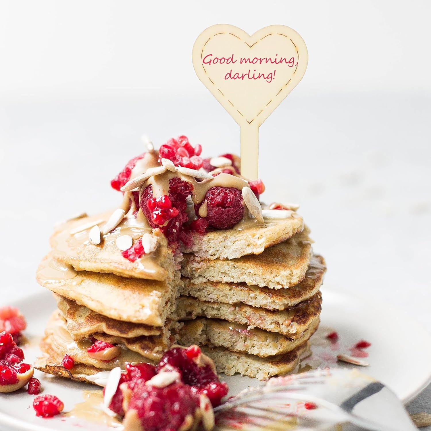 Stack of pancakes with raspberries and a heart-shaped topper on a white background