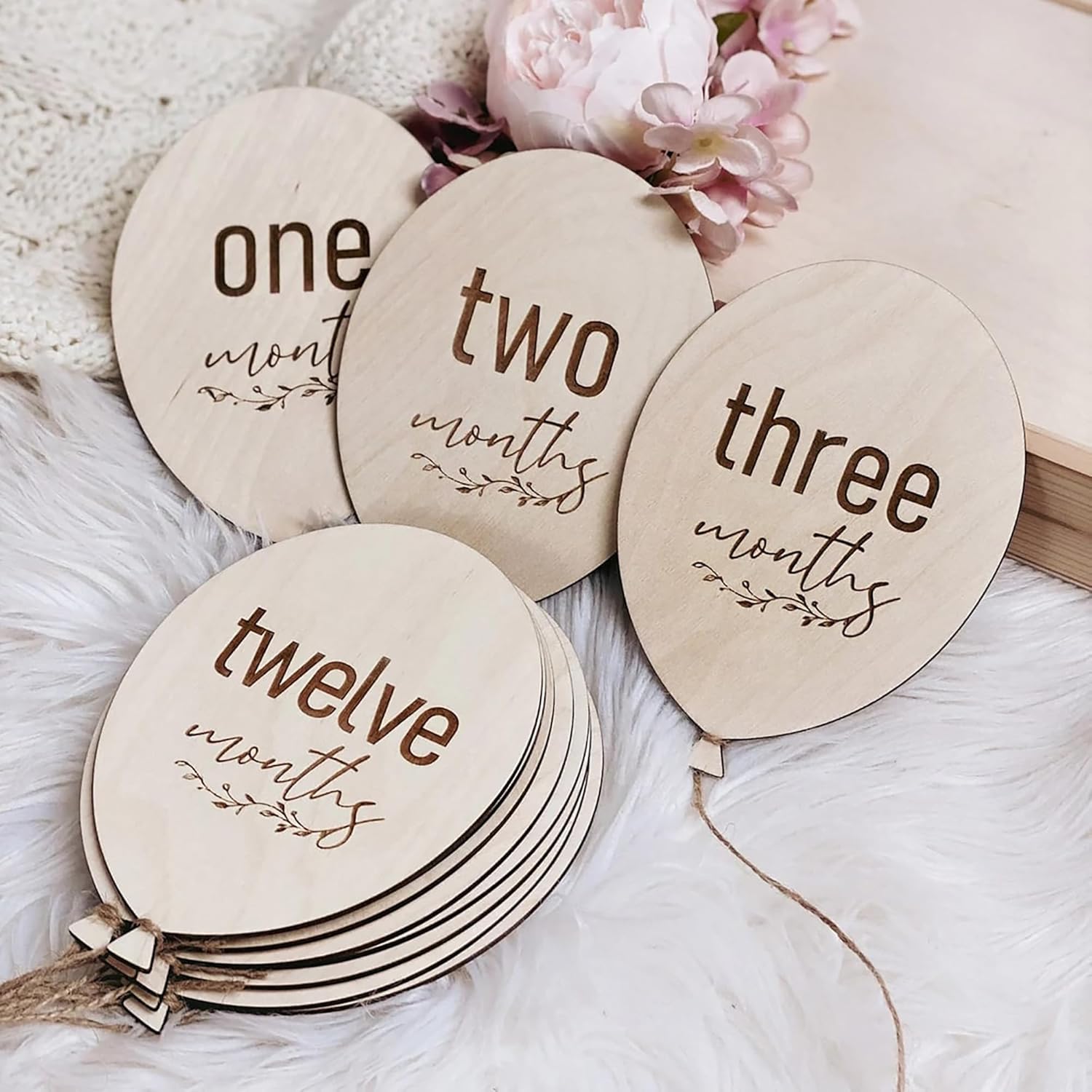 Wooden milestone markers for baby's first year with text 'one month', 'two months', 'three months', and 'twelve months' on a soft surface with flowers.