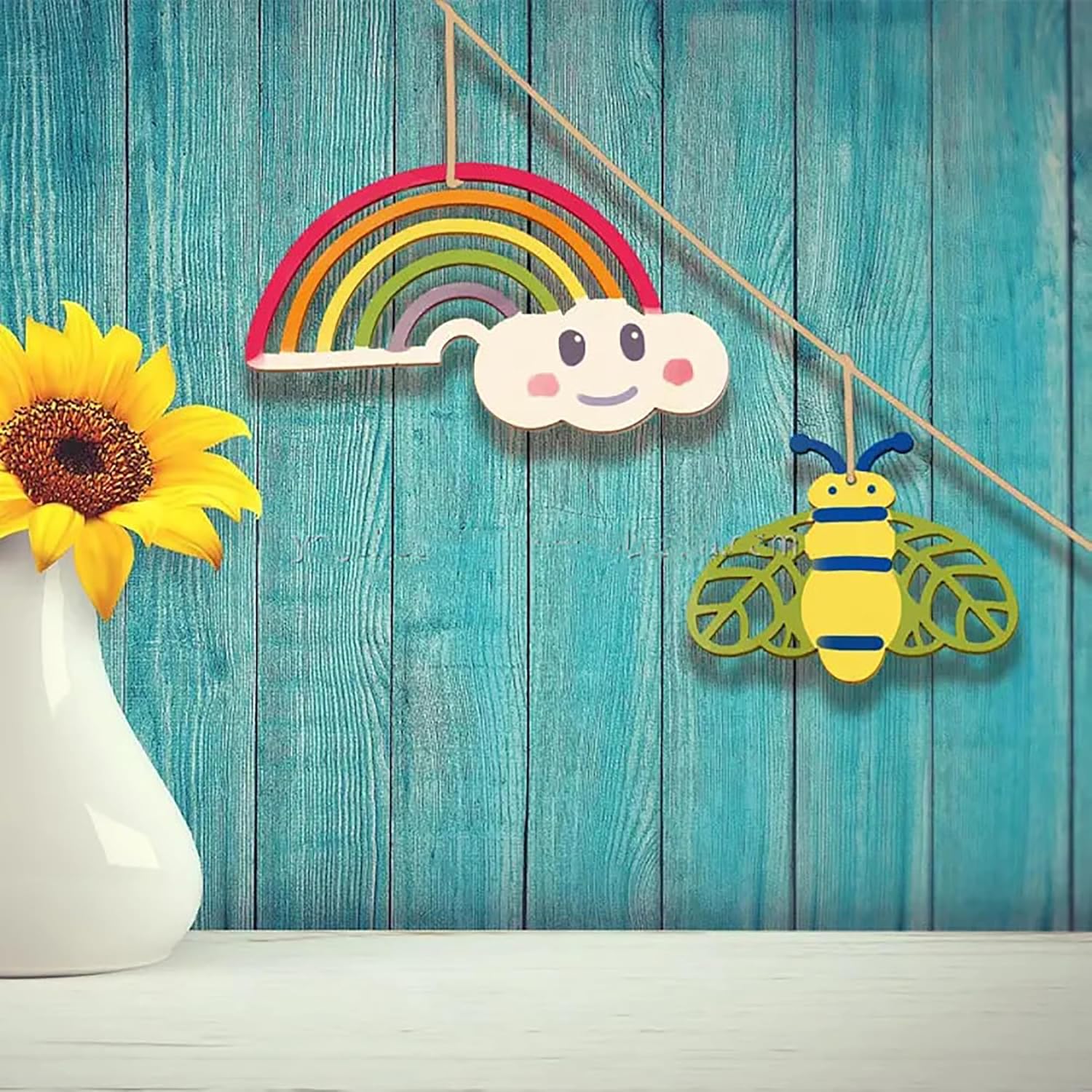 Decorative items including a rainbow, cloud, and bee on a teal wooden background with a sunflower in a white vase.