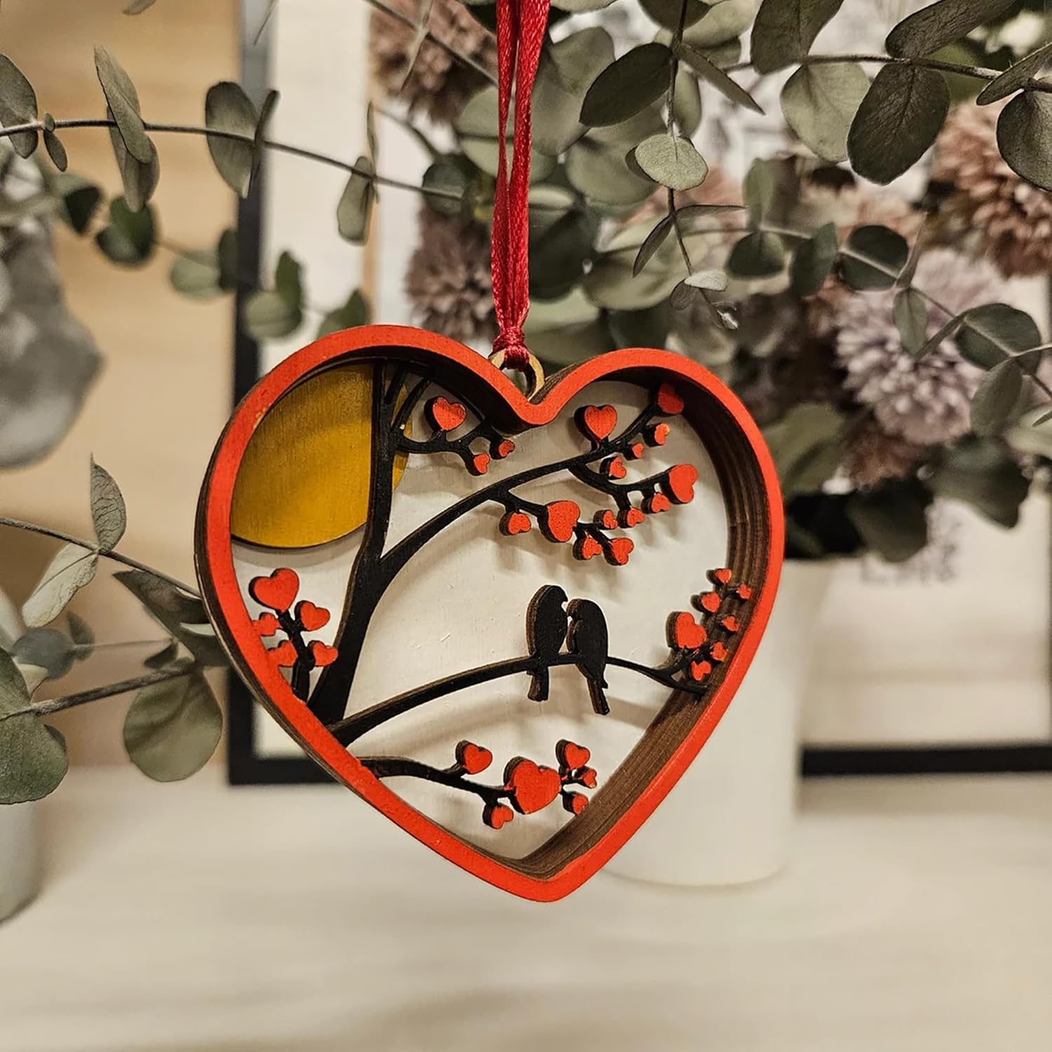 Heart-shaped ornament with a tree design and red hearts, hanging against a blurred natural background.