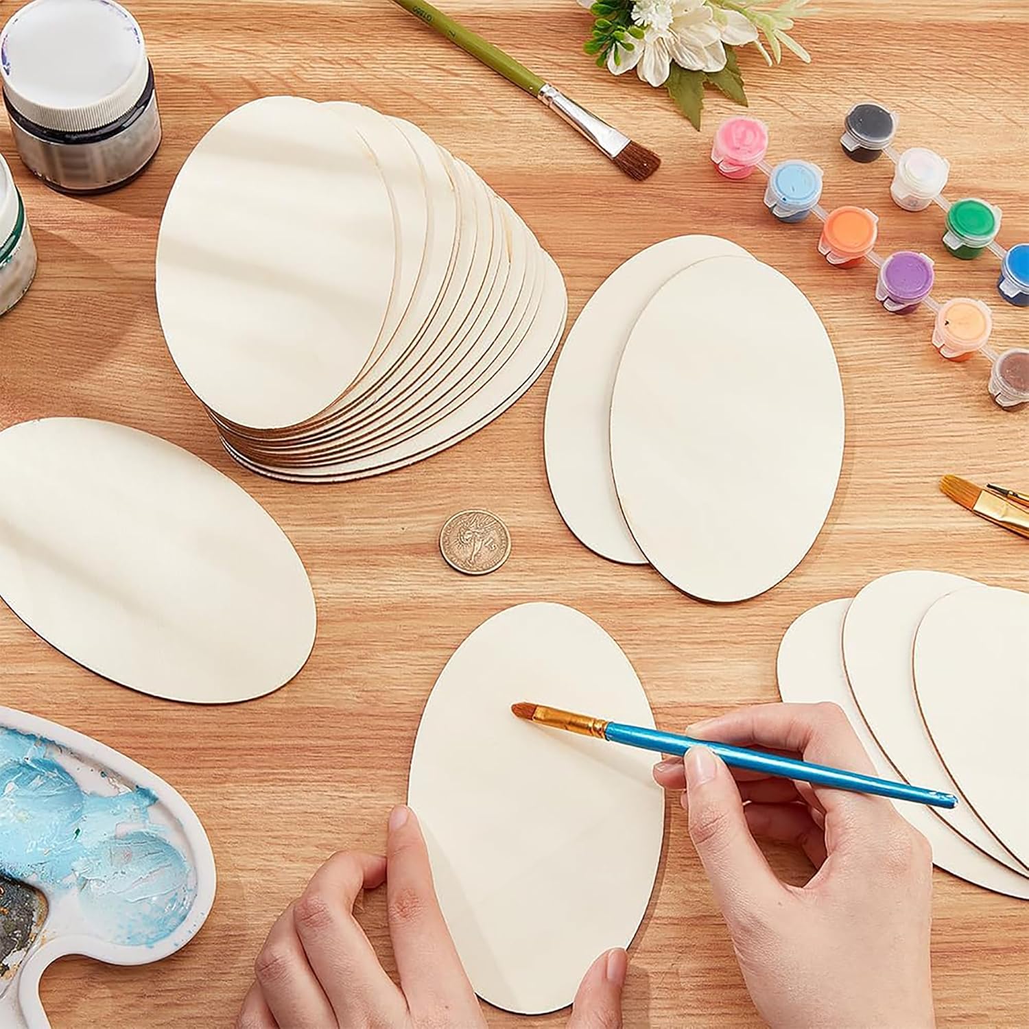 Hands painting oval-shaped wooden pieces on a wooden table with paint supplies.