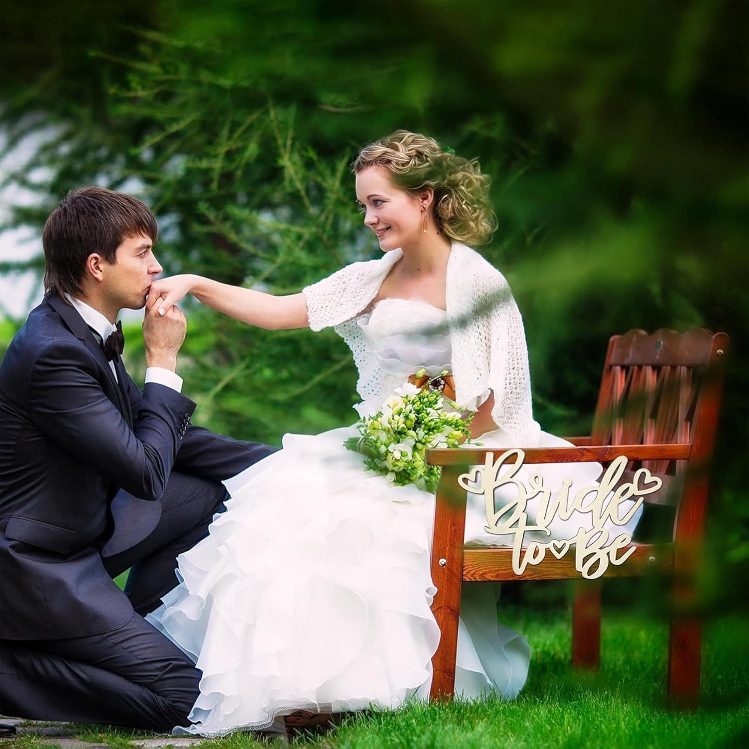 Couple in wedding attire sitting on a bench with 'Bride to be' sign in a garden setting.