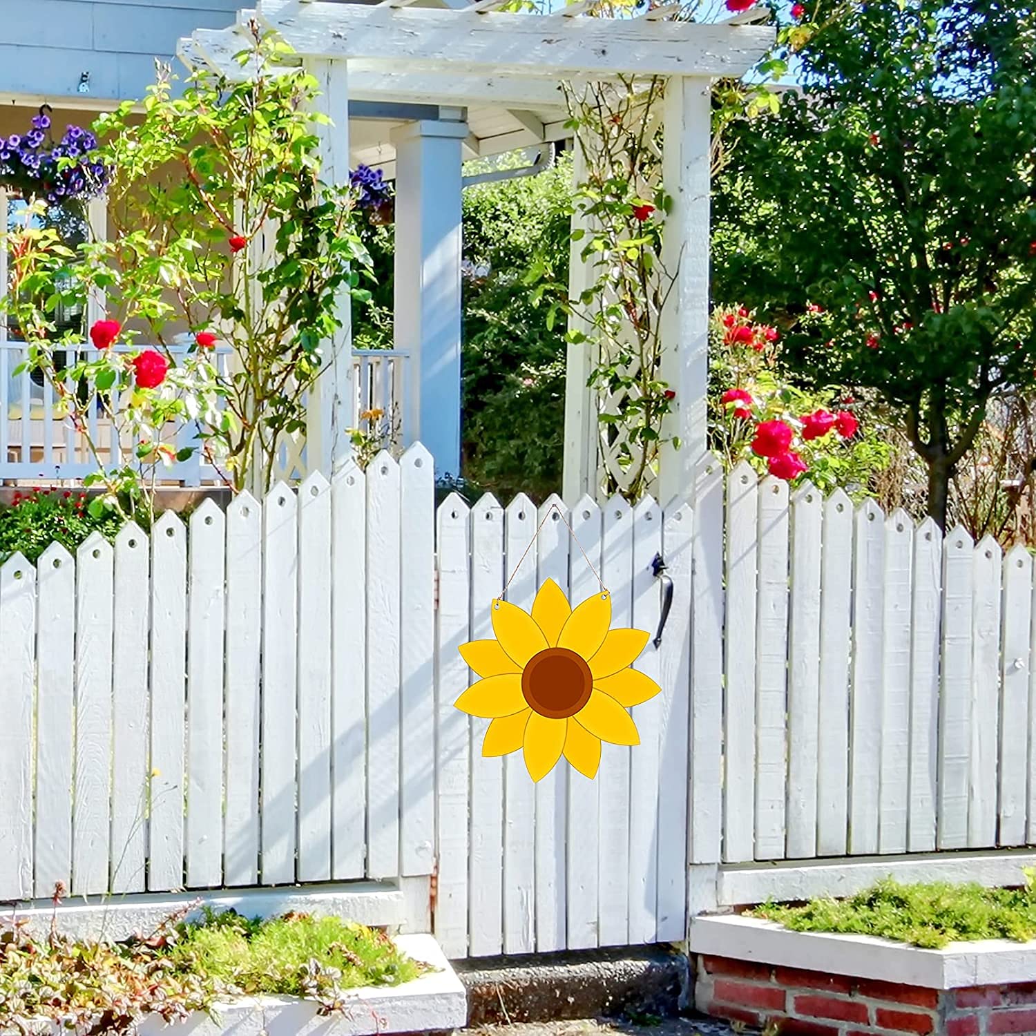 White picket fence with a sunflower decoration in front of a garden with flowers and a white arbor.
