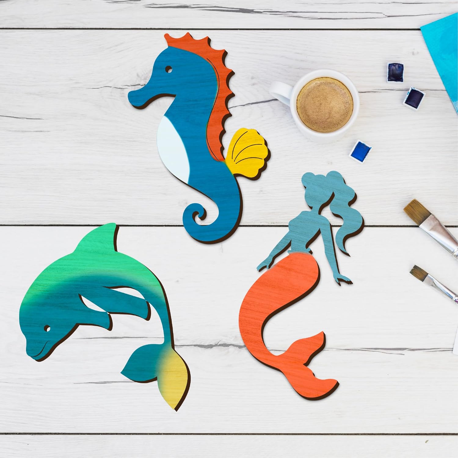 Colorful paper cutouts of a seahorse, dolphin, and mermaid on a wooden surface with paint and brushes.