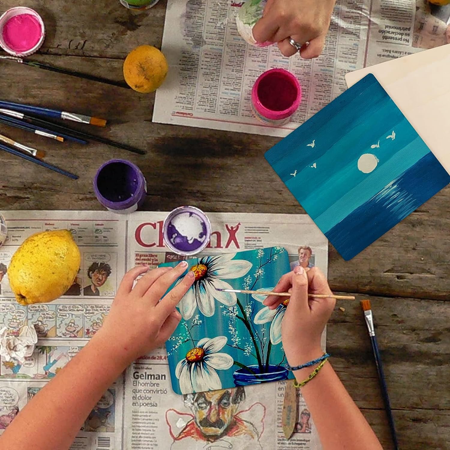 Person painting a floral design on a canvas with various art supplies on a wooden table.