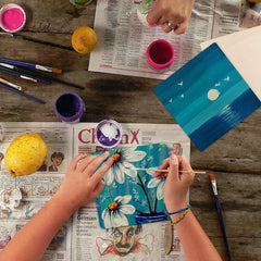 Person painting a floral design on a canvas with various art supplies on a wooden table.