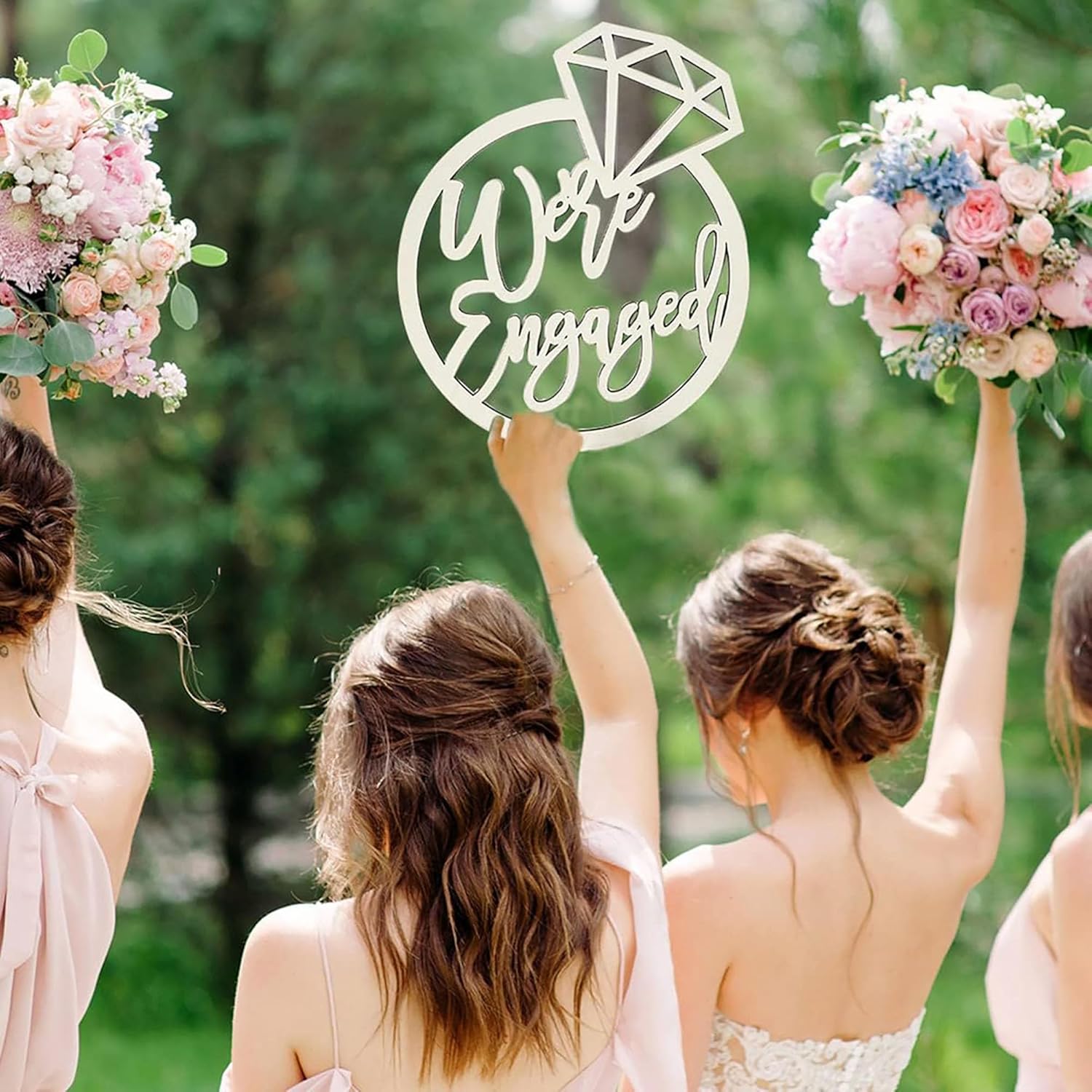 Four women holding flowers with a 'We're Engaged' sign in the center against a green outdoor background.