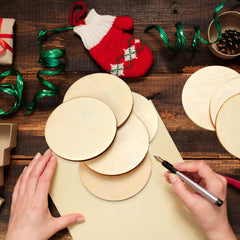 Hands working on a craft project with wooden circles and a pen on a wooden table, surrounded by Christmas decorations.