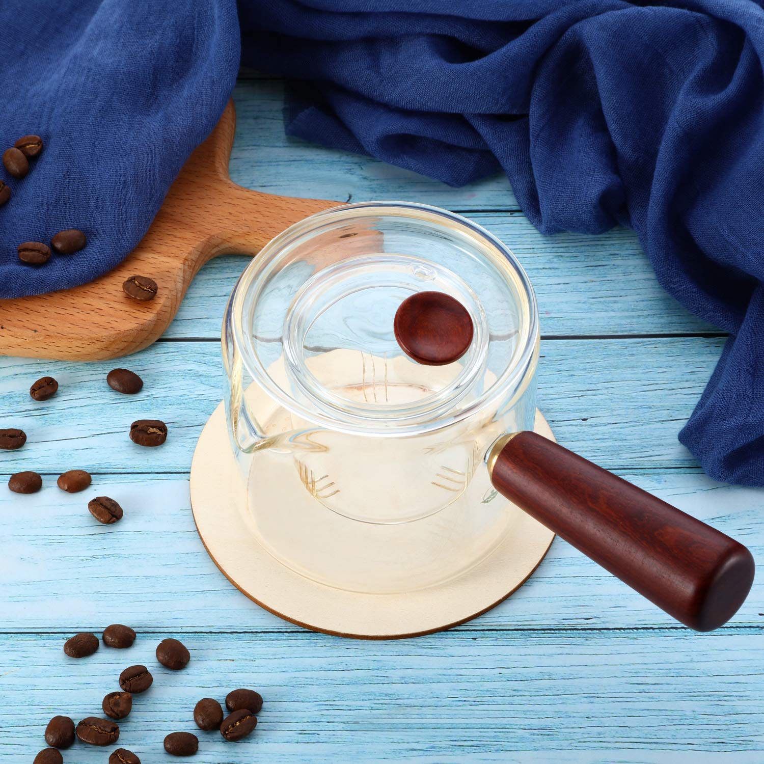 Glass coffee grinder with wooden handle on a blue wooden surface with scattered coffee beans.