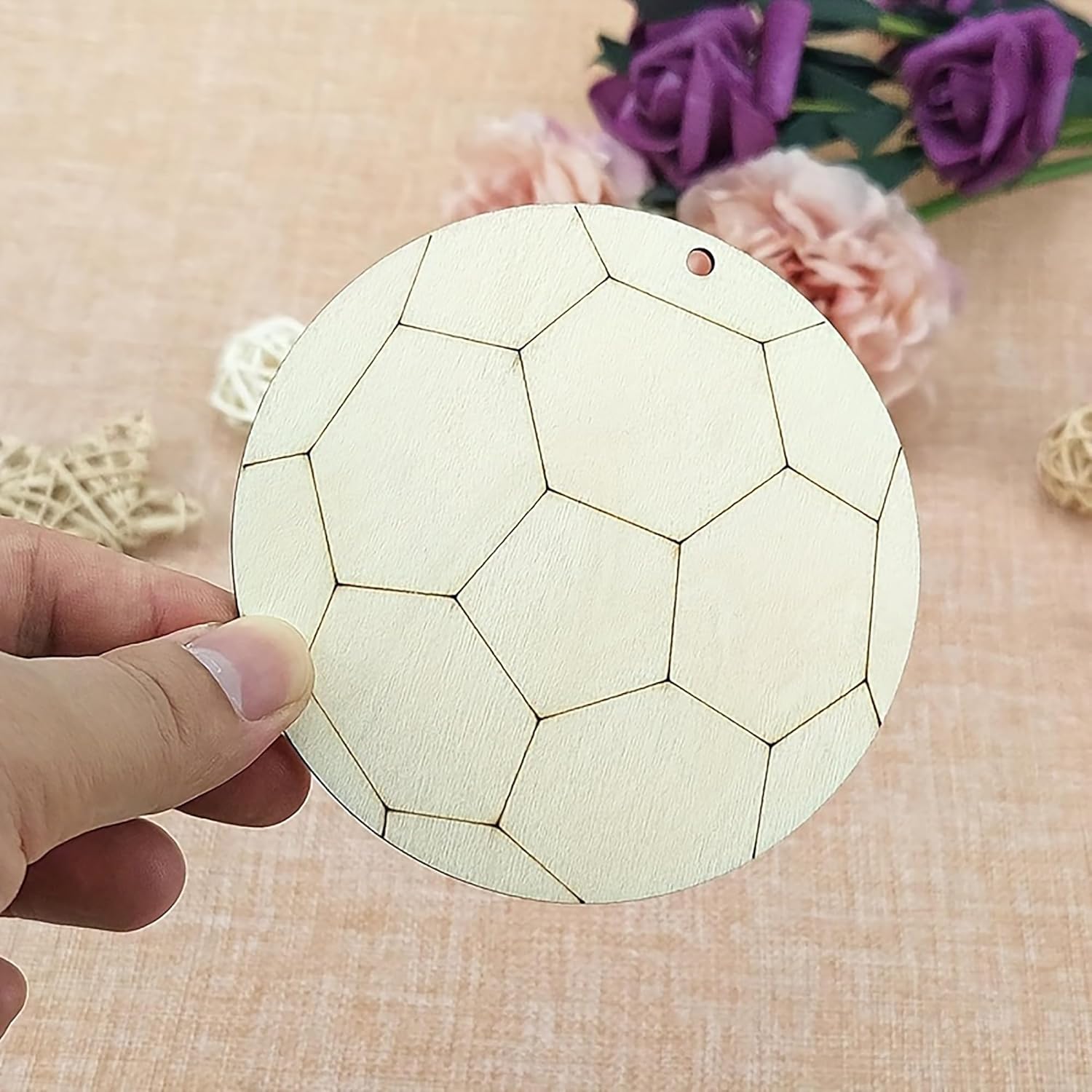 Hand holding a round wooden coaster with geometric pattern on a textured surface with flowers in the background
