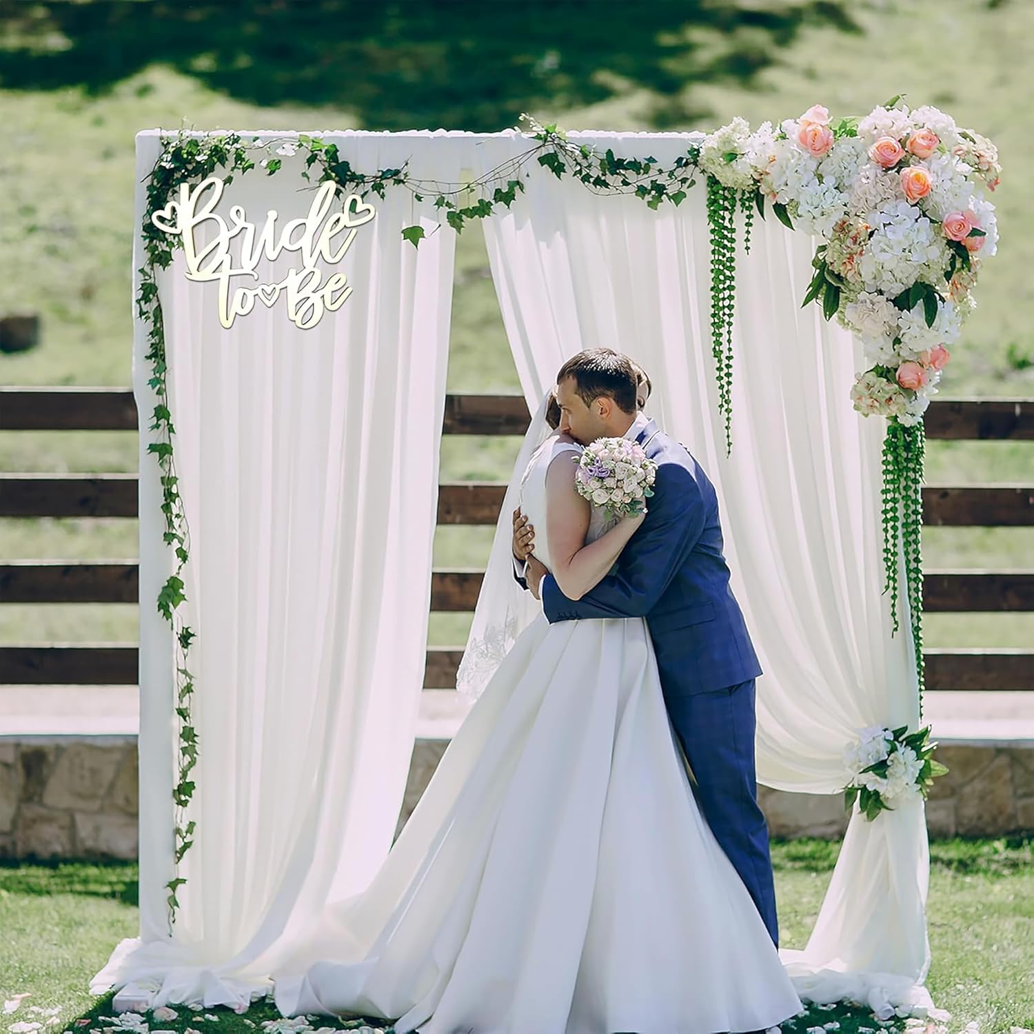 Couple embracing under a decorated arch with 'Bride to Be' sign, surrounded by greenery.