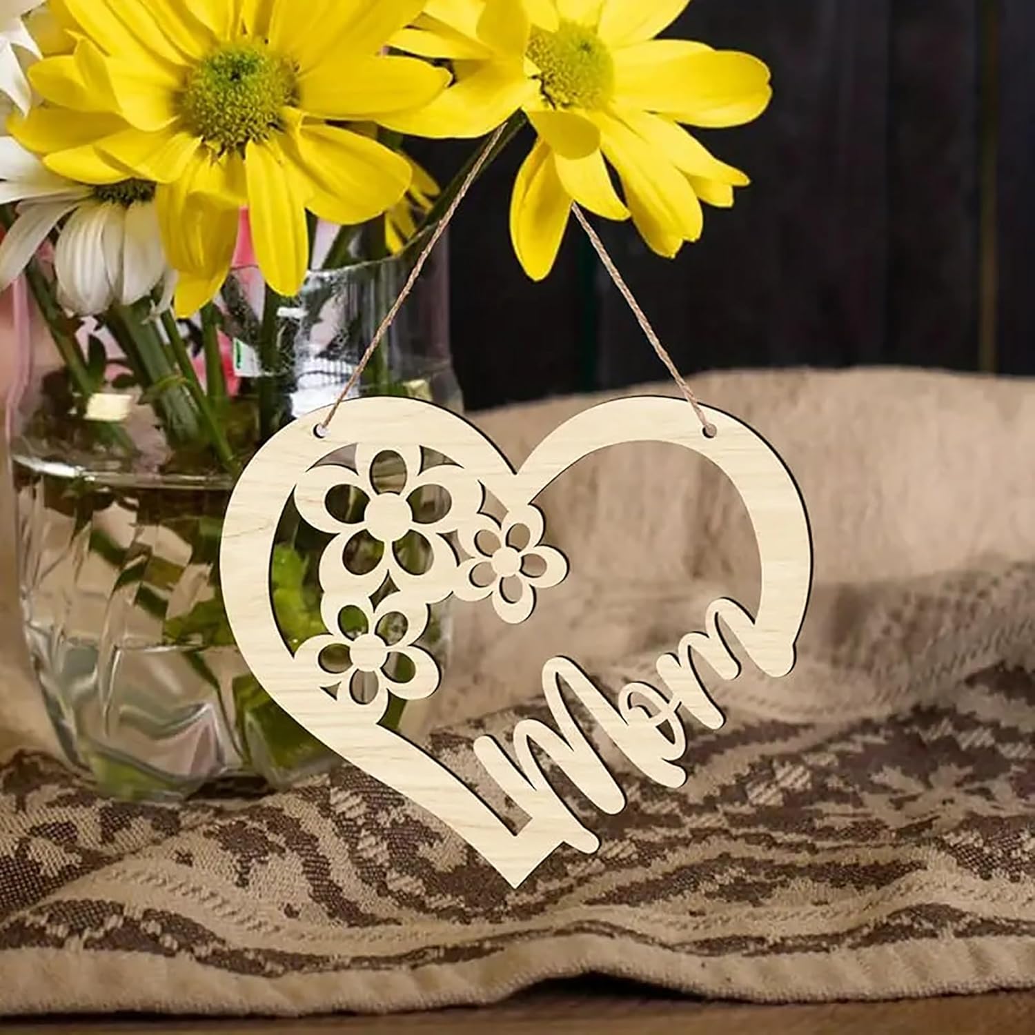 Heart-shaped wooden ornament with 'Mom' engraved, placed in front of a vase with yellow flowers.