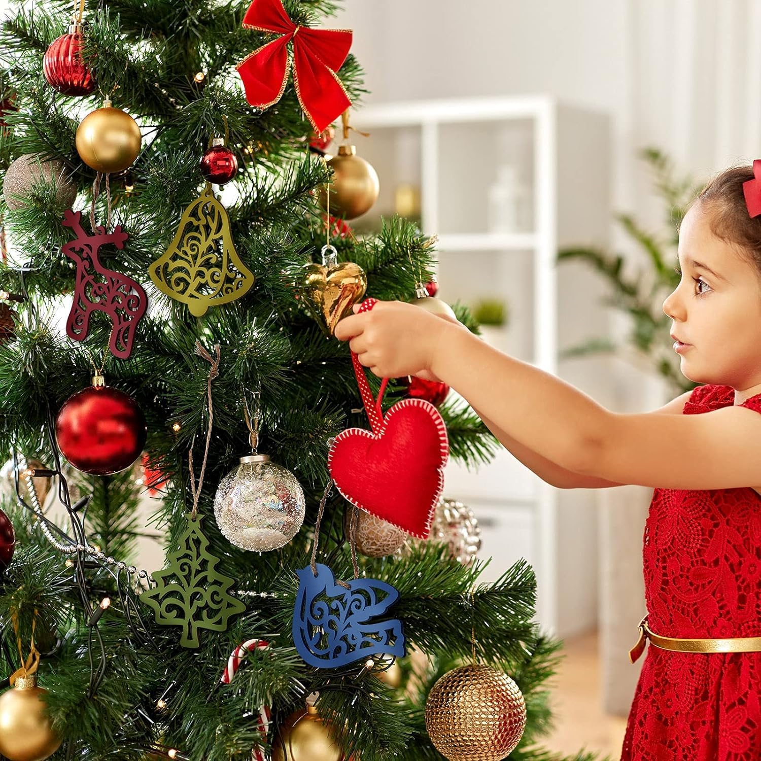 Child in a red dress decorating a Christmas tree with various ornaments.