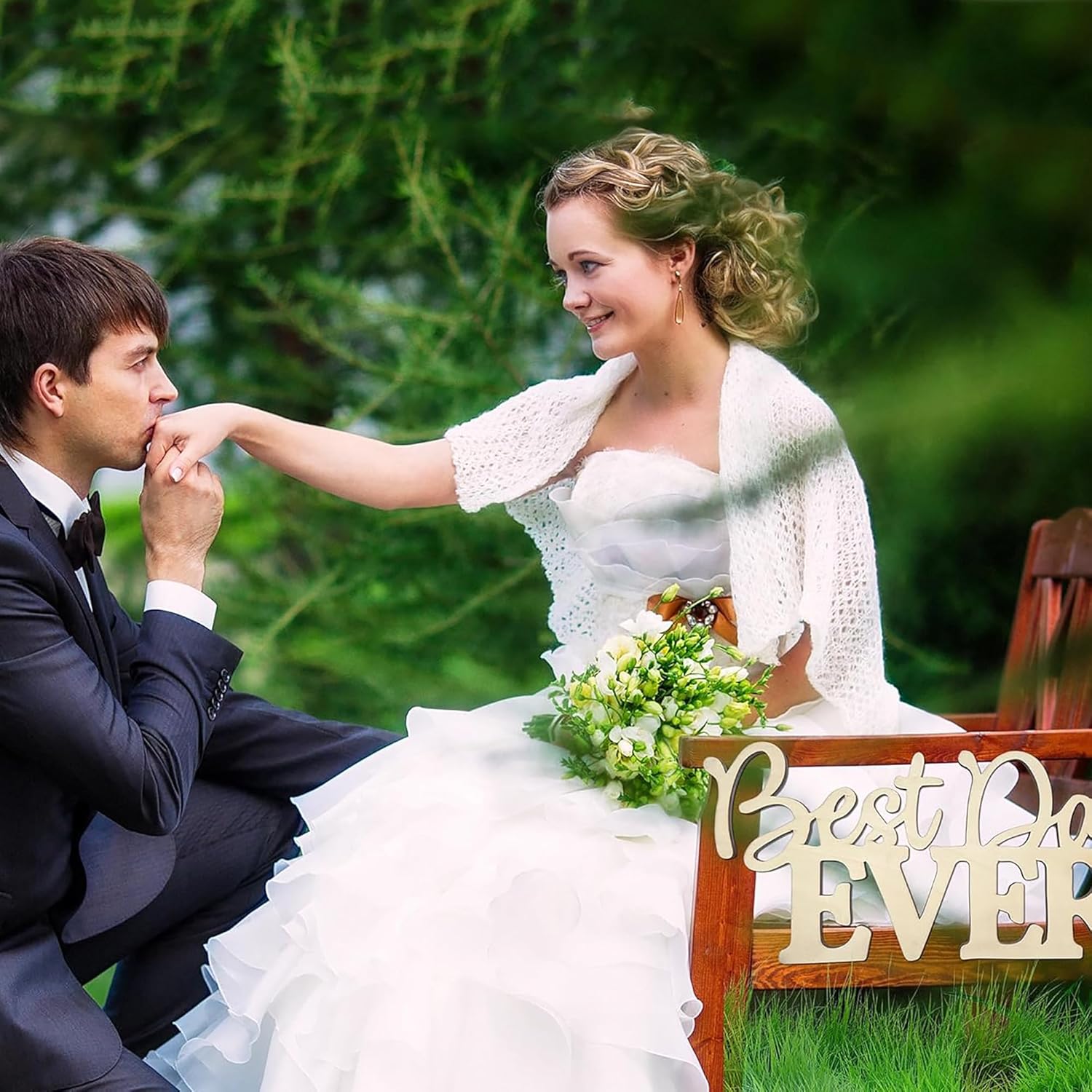 Couple in wedding attire sitting on a bench with 'Best Day Ever' sign in a garden setting.