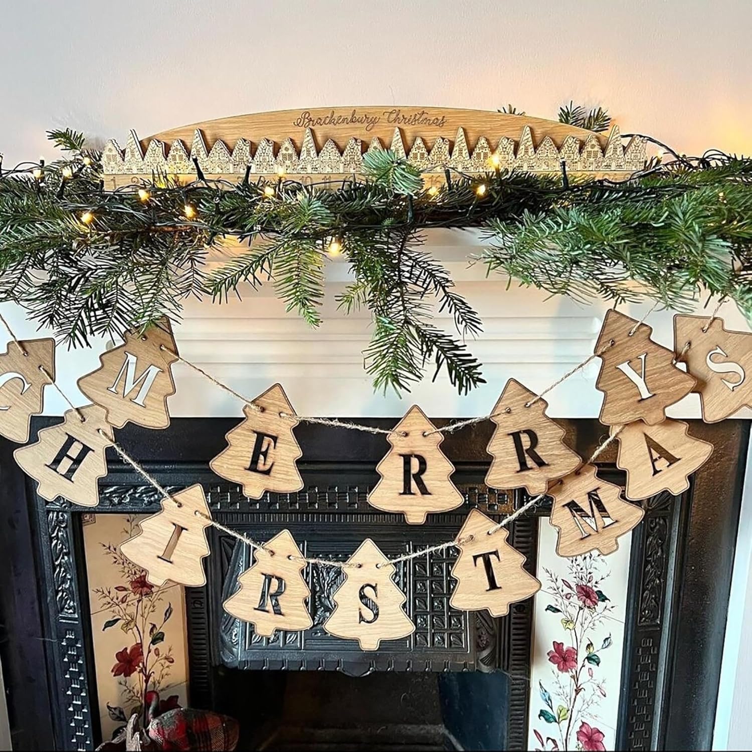 Wooden 'Merry Christmas' banner on a decorated fireplace mantel with greenery and lights.