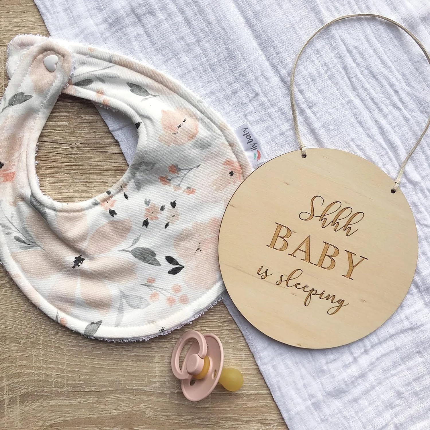 Baby bib with floral pattern, pacifier, and 'Shh Baby is sleeping' sign on a textured surface.