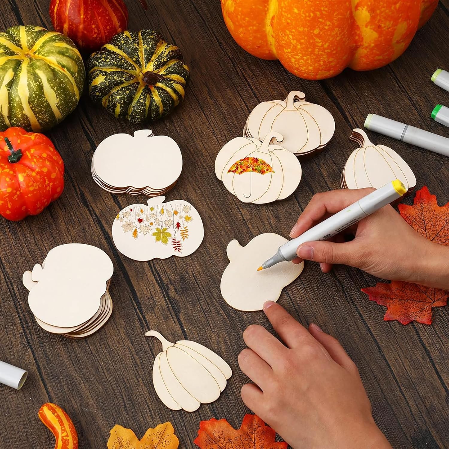 Wooden pumpkin-shaped cutouts with markers on a wooden surface with pumpkins and leaves.