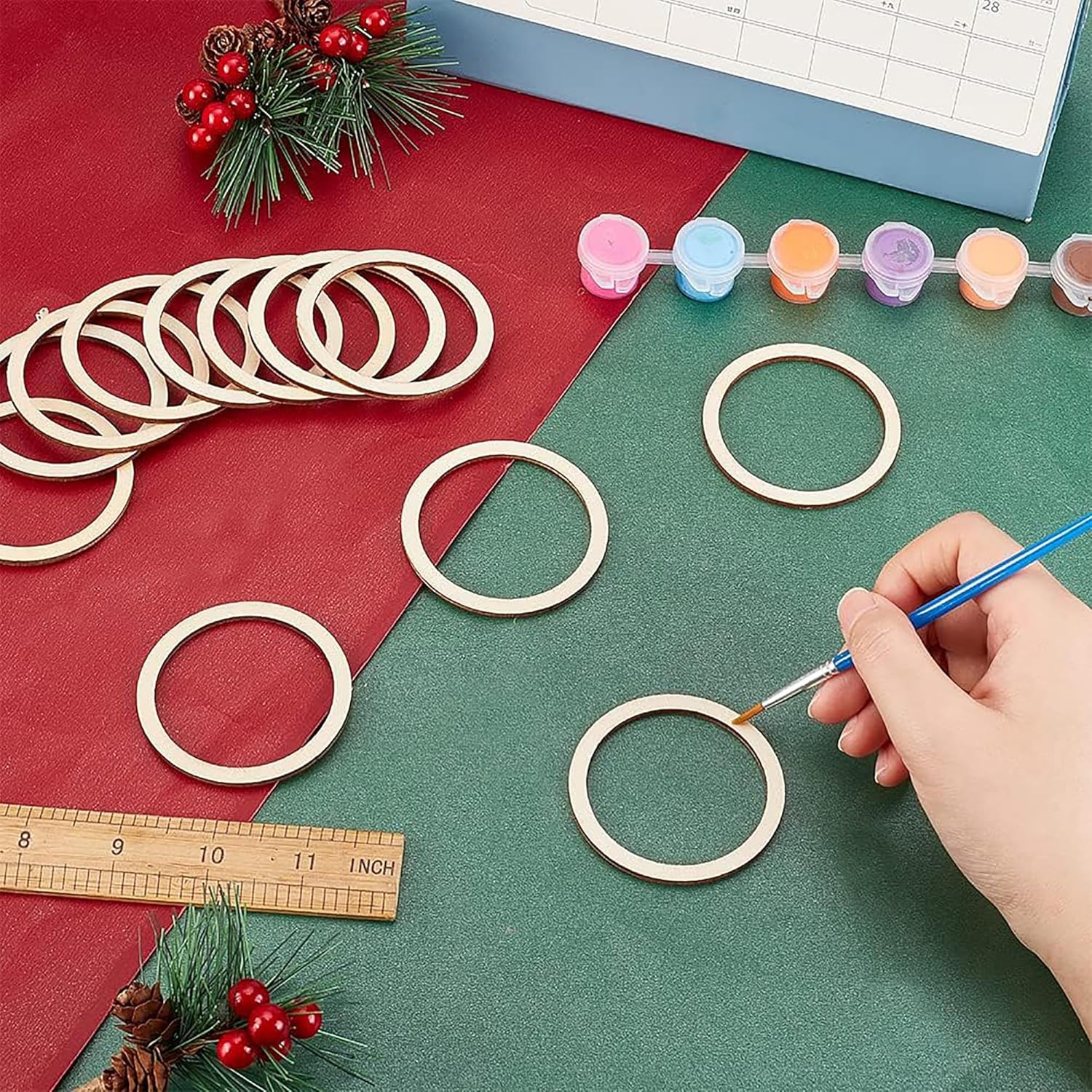 Hand painting wooden rings on a green surface with Christmas decorations and tools.
