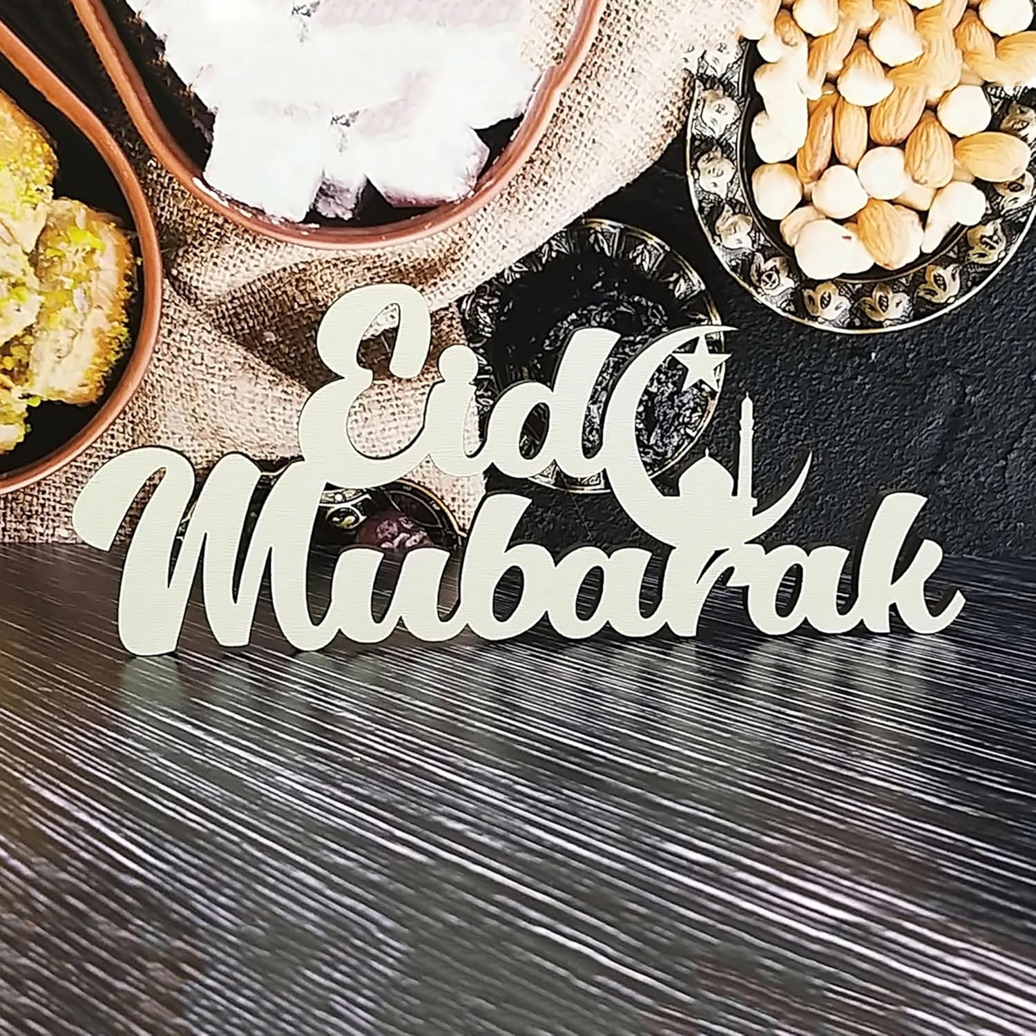 Wooden 'Eid Mubarak' sign on a dark surface with food items in the background