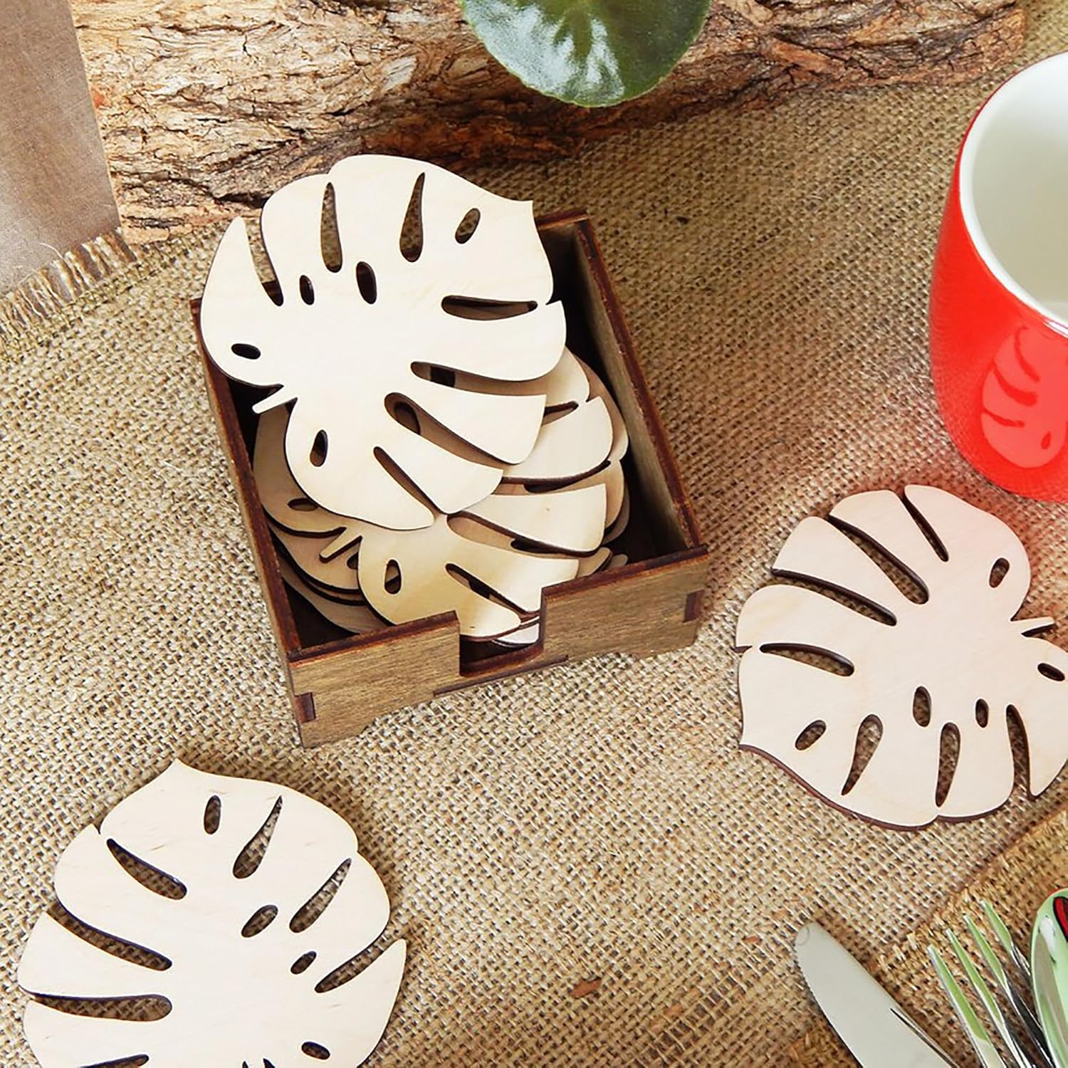 Wooden leaf coasters in a box on a textured surface with a red mug and green plant.