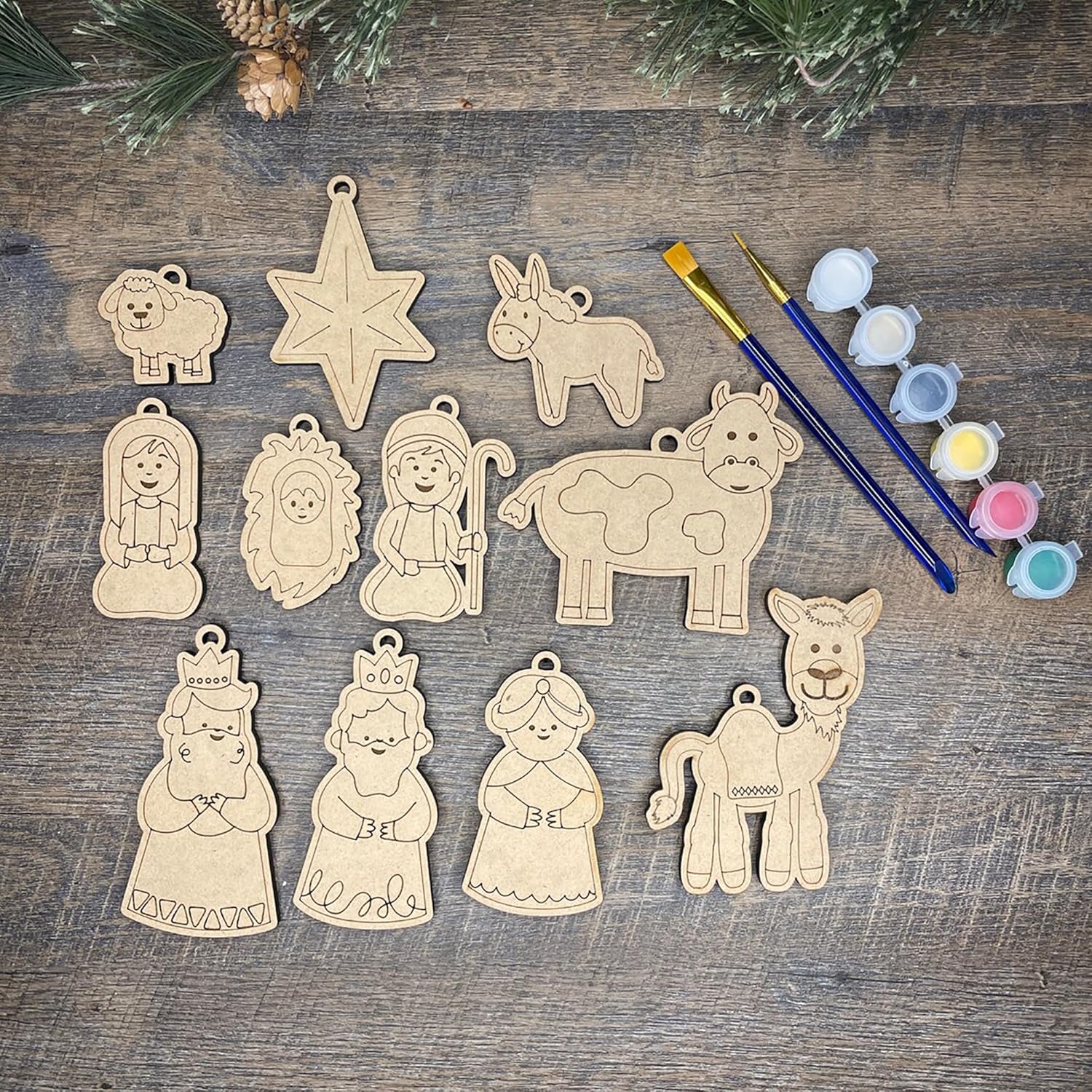 Set of wooden nativity scene ornaments on a wooden surface with paintbrushes and paint.