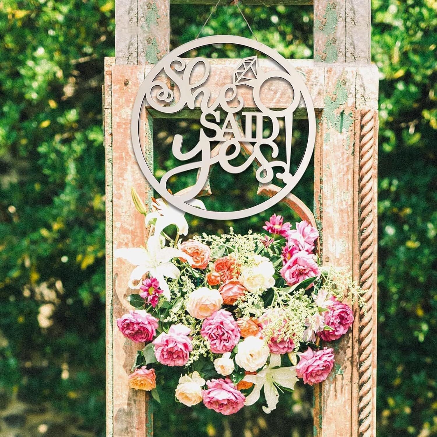 Decorative sign with 'She Said Yes' surrounded by flowers on a wooden stand against a green foliage background