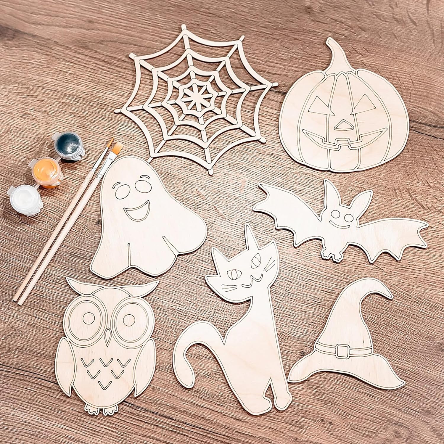 Halloween-themed wooden cutouts including a ghost, spider web, pumpkin, bat, cat, owl, and witch hat on a wooden surface with paint and brushes.
