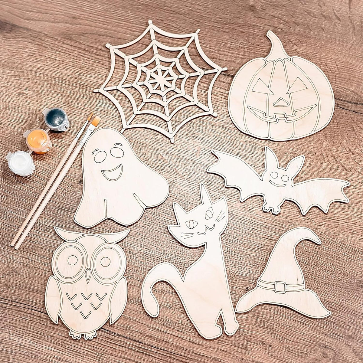 Halloween-themed wooden cutouts including a ghost, spider web, pumpkin, bat, cat, owl, and witch hat on a wooden surface with paint and brushes.