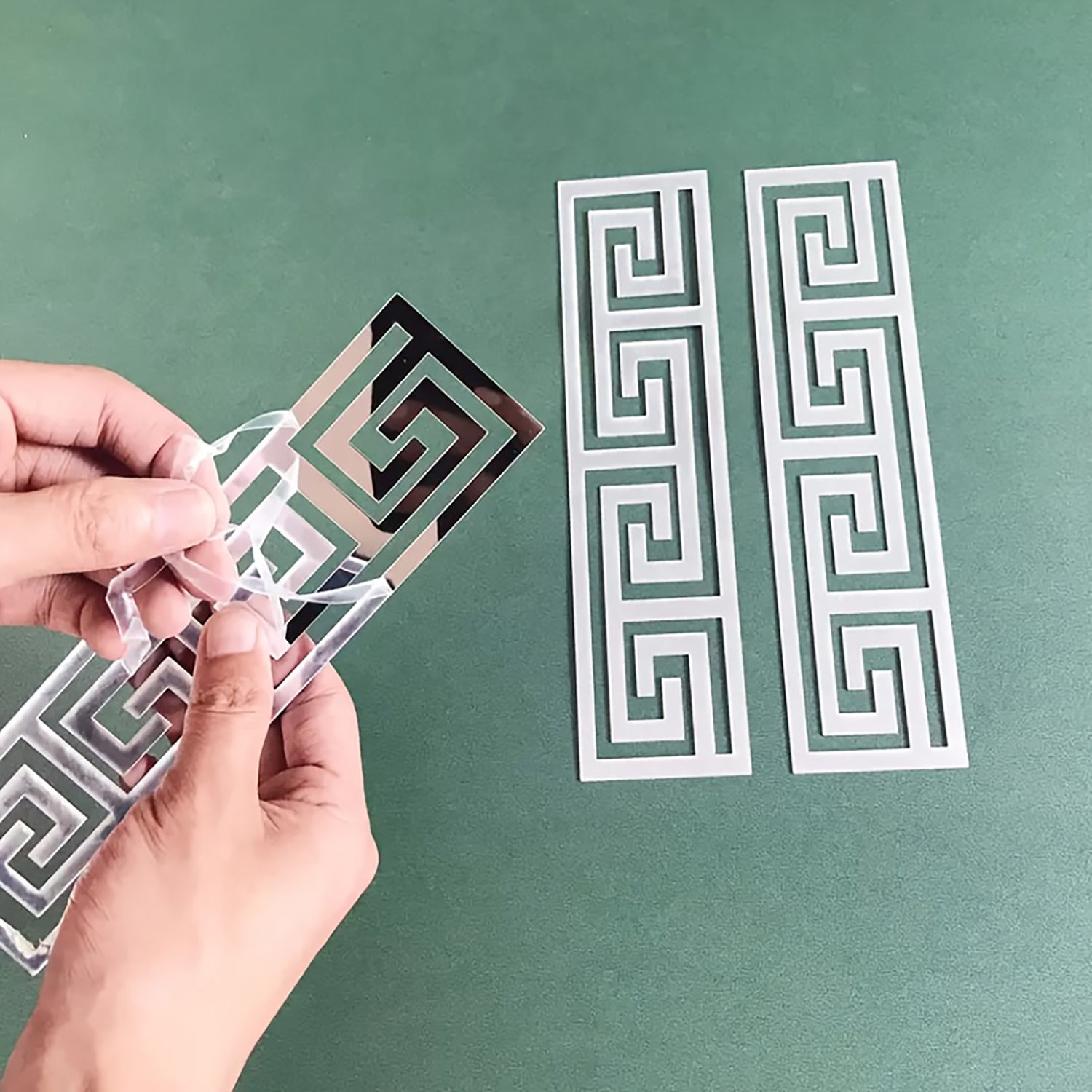 Hand holding a metallic stencil with Greek key pattern on a green background