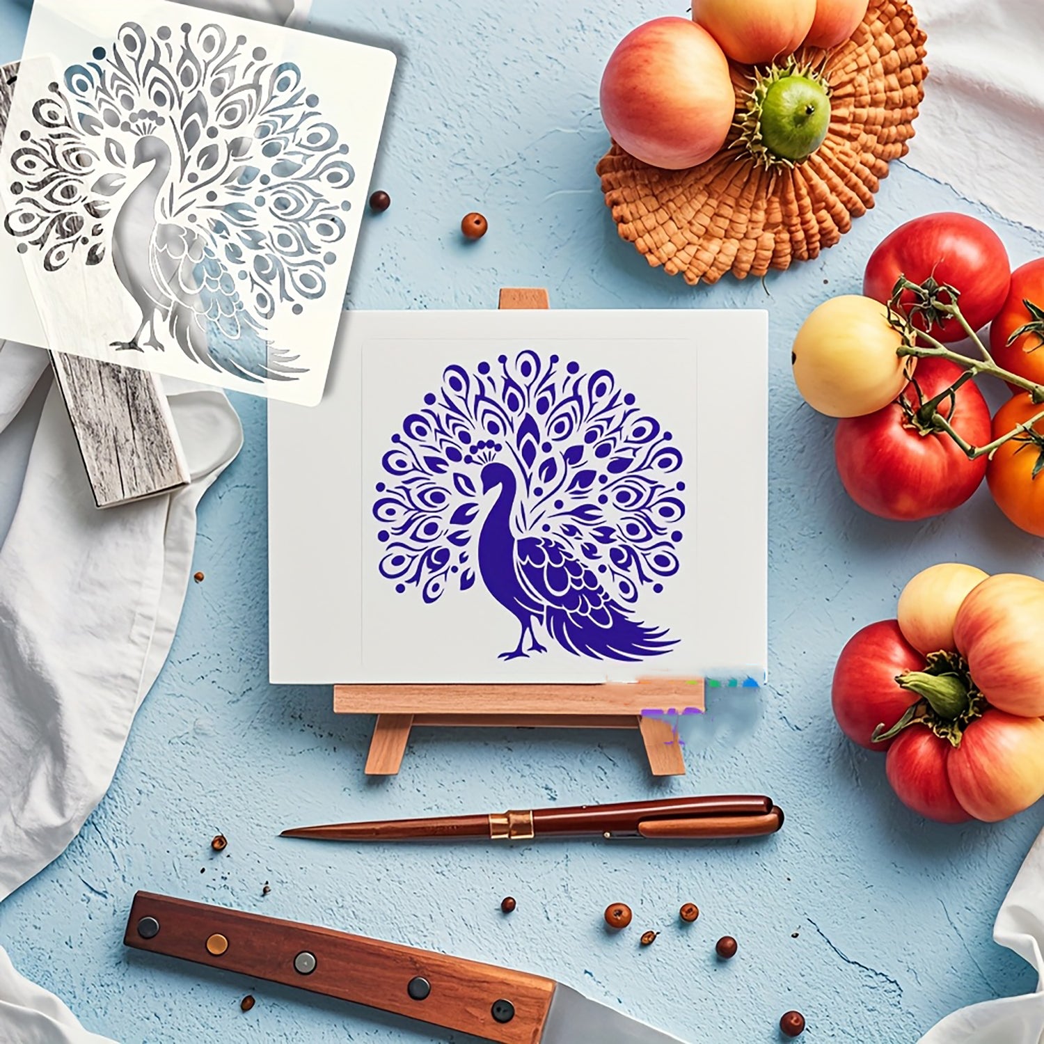 Peacock design on a small canvas with a knife, tomatoes, and peaches on a blue surface