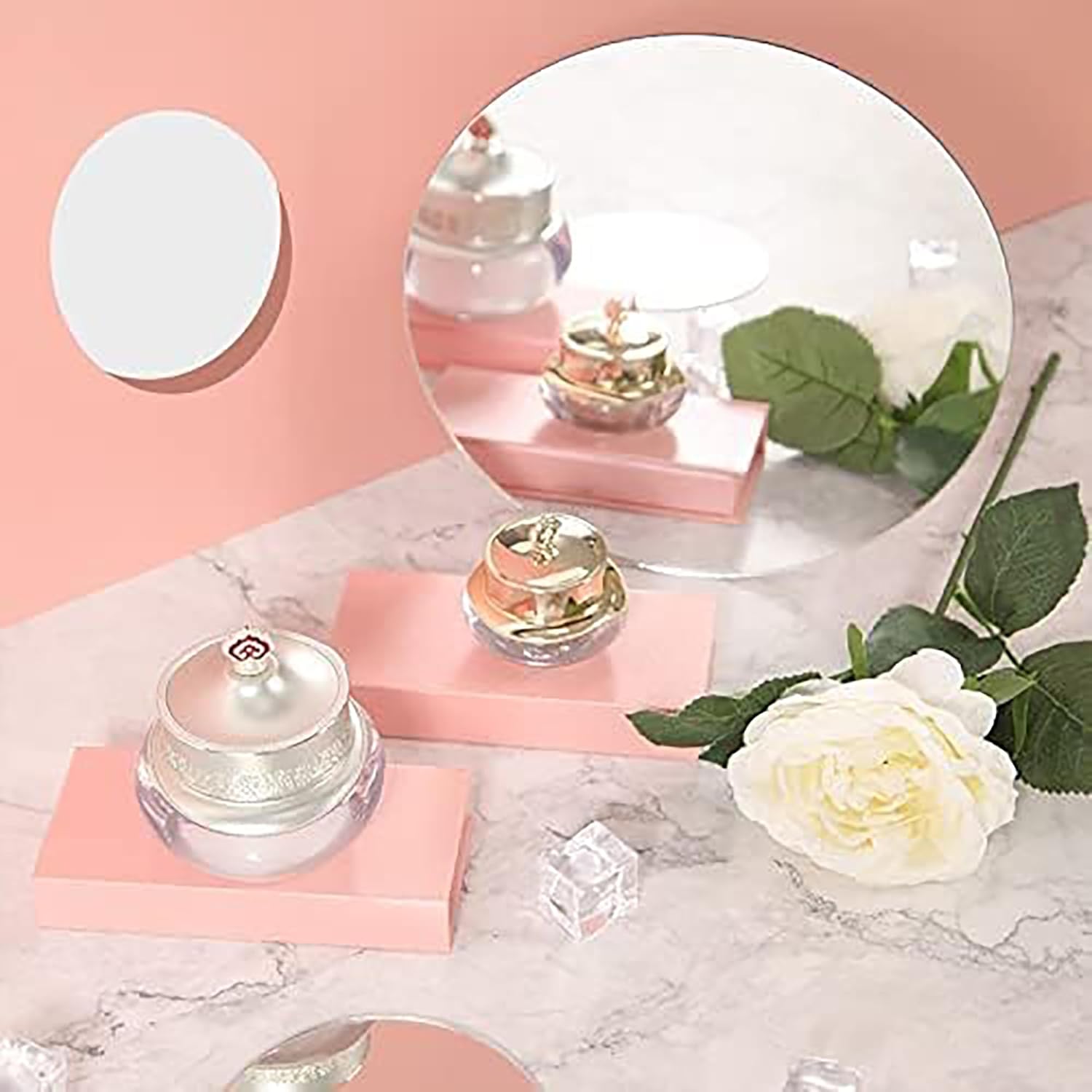 Cosmetic jars on pink stands with a white rose and green leaves on a marble surface.